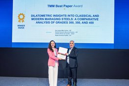 Man hands over a certificate to a woman. They are standing on a stage. A presentation is shown in the background.