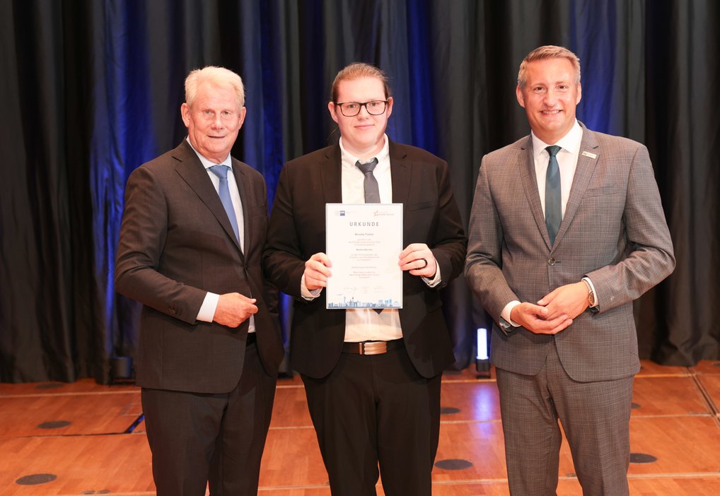 Three men in suits are standing next to each other. The younger man in the middle is holding a certificate.