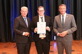 Three men in suits are standing next to each other. The younger man in the middle is holding a certificate.