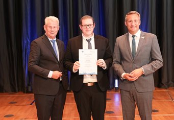 Nicolas Fiebes (centre) was honoured by the Düsseldorf Chamber of Industry and Commerce (IHK) as one of the top graduates of its 2026 vocational training and advanced professional qualification examinations. He received his certificate from Düsseldorf District President Thomas Schürmann (right) and IHK President Andreas Schmitz (left). Three men in suits are standing next to each other. The younger man in the middle is holding a certificate.